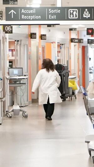 Une femme vêtue d'un sarrau blanc marche dans un corridor d'hôpital où l'on voit une civière.