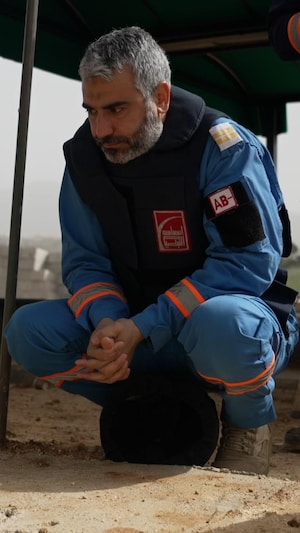 Un homme aux cheveux gris est accroupi près d'une tombe. Il est en habit de secouriste bleu, un casque est à ses pieds. Il porte un gilet pare-balles.