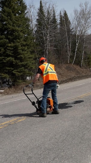 Un ouvrier en vêtements de sécurité colmate un trou dans la chaussée. Un camion qui transporte des cônes orange est stationné en bordure de route. 