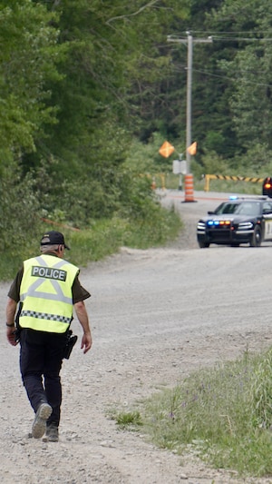 Un policier marche dans un chemin boisé. Deux voitures de police sont stationnées plus loin.