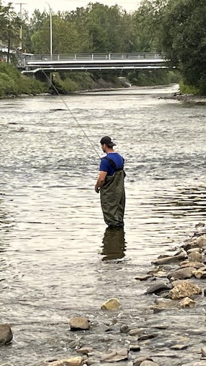Un homme pêche debout dans une rivière, tandis qu'on voit un petit pont au loin.