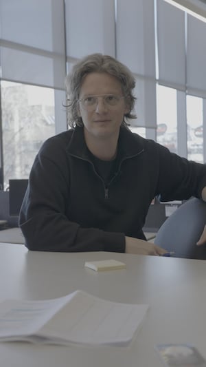 Pierre-Yves McSween est assis à une table, il regarde devant lui, une journaliste lui pose des questions.