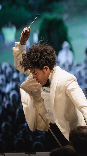 Le chef d'orchestre en pleine action, baguette dans les airs, avec une foule diffuse en arrière-plan. 