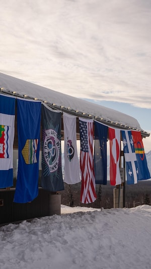Les huit drapeaux des délégations suspendus les uns à côté des autres, en fond une chaîne de montagnes, aux Jeux de l'hiver de l'Arctique 2024, à Mat-Su en Alaska. 