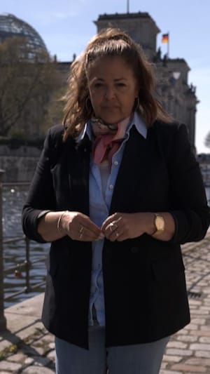 La journaliste Tamara Altéresco est debout, sur un trottoir pavé de Berlin. Elle se trouve près de l'eau. Derrière elle, on voit le parlement allemand. 