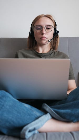Une jeune femme portant des lunettes et un casque d'écoute est assise sur un sofa devant son ordinateur portable ouvert.