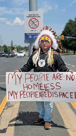 Le chef Hosea Wesley porte un chapeau traditionnel autochtone avec des plumes et présente une pancarte avec inscrit « mon peuple n'est pas sans-abri » en anglais.
