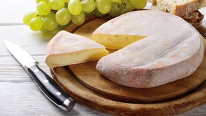 Photo d'un reblochon sur une planche en bois avec une tranche coupée. Un couteau est posé à côté et on aperçoit du pain et des raisins en arrière plan.