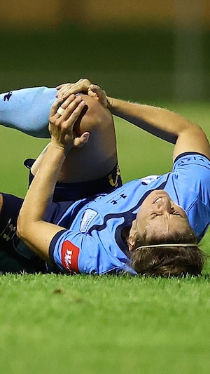 Une joueuse tient son genou droit entre ses mains, couchée sur son dos en douleur sur la pelouse d'un terrain de soccer.