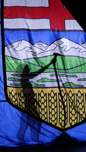 Une personne vue seulement en silhouette défroisse un drapeau albertain de très grande ampleur. 