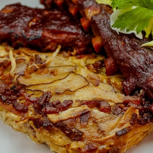 Un gratin de panais, pommes de terre rattes et bacon qui accompagne les côtes levées dans une assiette.