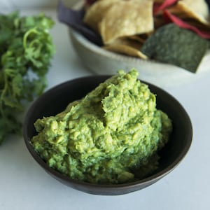 Un bol de purée d'avocat à côté de croustilles de ma¨s et d'un bouquet de coriandre.