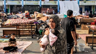 Une mère tenant un bébé marche au milieu de lits et de personnes dans le désordre d'un camp de réfugiés.