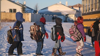 Quatre enfants habillés pour l’hiver avec des sacs à dos font la file devant un autobus scolaire jaune.