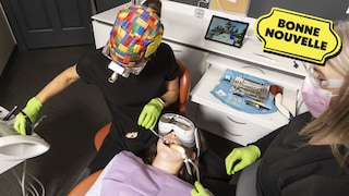 Une jeune fille est assise dans une chaise et porte un casque de réalité virtuelle. Deux professionnelles de la santé dentaire inspectent sa bouche.