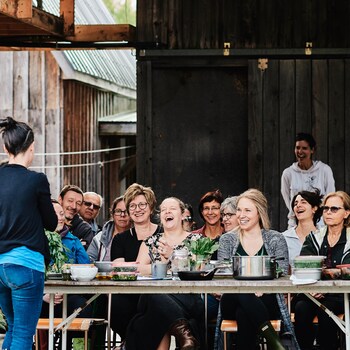 Une personne donne un atelier culinaire devant un groupe de gens assis derrière de longues tables en bois. 