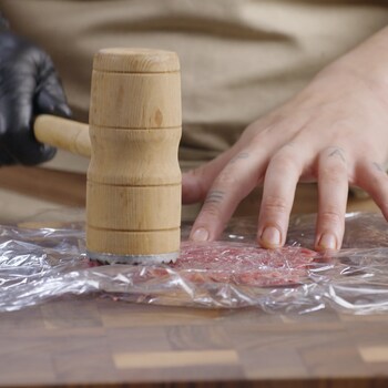 Une personne utilise le côté plat du maillet pour aplanir le morceau de viande sur une surface de travail.
