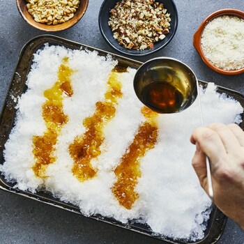 Un contenant rempli de neige sur une table avec différentes garnitures à l'entour. Une main verse de la tire d'érable sur la neige.