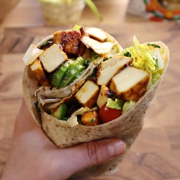 Une main tenant un wrap aux bâtonnets de tofu magique au BBQ et aux légumes, avec des ingrédients sur une table en bois en arrière-plan.