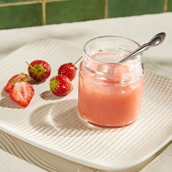 Un pot de verre contenant du gel de fraises accompagné de quelques fraises fraîches.