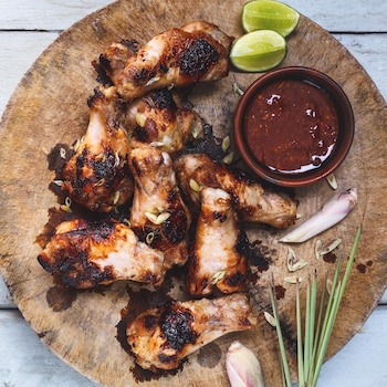 Des ailes de poulet grillées avec un bol de sauce et des quartiers de lime sur une plaque en bois.