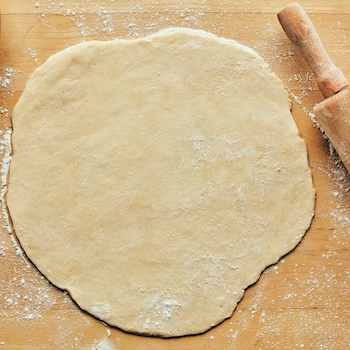Sur un comptoir, est déposé la pâte à tarte, un rouleau pour abaisser la pâte et une assiette. 