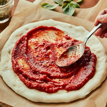 Une main étend la sauce à pizza sur une pâte à pizza non cuite avec une cuillère. 