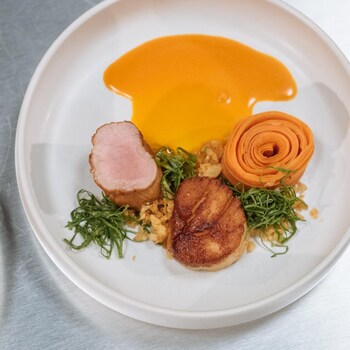 Dans une assiette se trouve un médaillon de filet de porc, un pétoncle grillé et une rosace de carottes. Le tout est arrosé d'une sauce orangée.