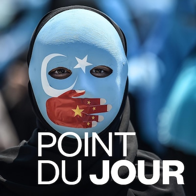 Une manifestante porte un masque aux couleurs du drapeau du Turkestan oriental. Une main représentant le drapeau chinois est peinte et couvre sa bouche. On ne voit que les yeux.