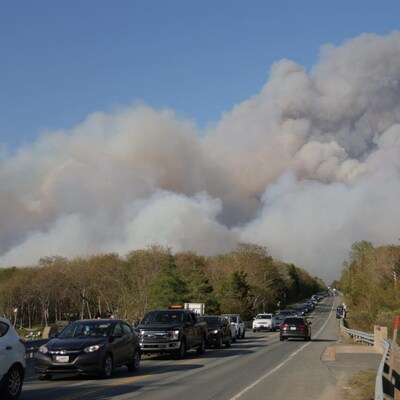 Une file de voitures en rang sur l'autoroute près de l'affiche qui souhaite la bienvenue à Stillwater Lake. Une épaisse fumée de feu de forêt couvre le ciel.