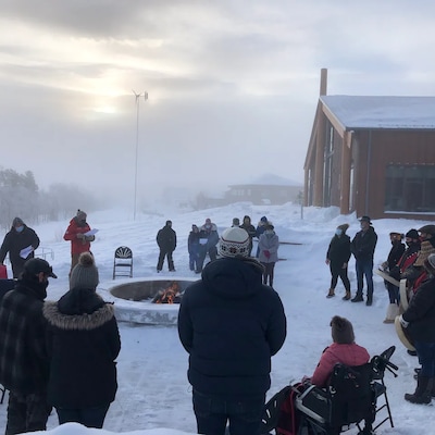 Une trentaine de personnes sont réunies autour d'un feu de camp et entourées de neige près d'une maison.