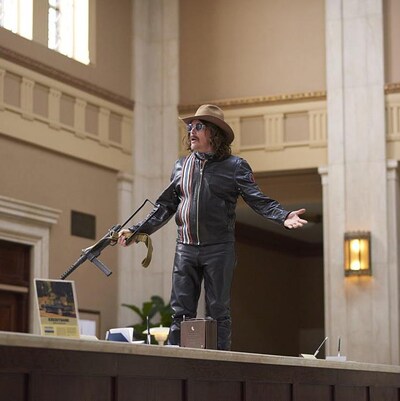Un homme habillé de cuir, un chapeau de cowboy et tenant une mitraillette est debout sur le comptoir d'une banque.