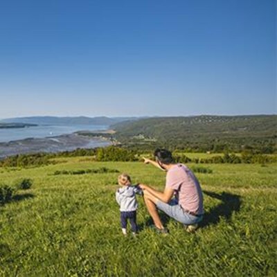 Un bébé et son père sont dans un dans un champ vallonné sur le bord du fleuve Saint-Laurent. Le père pointe vers l'horizon.