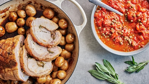 Un rôti de porc et des pommes de terre grelots dans une casserole sortant du four et un bol avec une sauce aux tomates cerises confites.