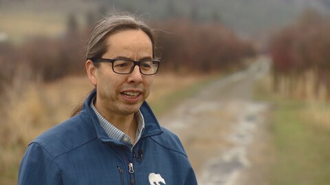 Le chef de la communauté de Lower Nicola, Aaron Sumexheltza, porte des lunettes noires et un manteau bleu. Il se dresse devant une route mouillée par la pluie et bordée d'arbres sur lesquels les feuilles peinent encore à verdir. Le chef de la communauté de Lower Nicola, Aaron Sumexheltza, porte des lunettes noires et un manteau bleu. Il se dresse devant une route mouillée par la pluie et bordée d'arbres sur lesquels les feuilles peinent encore à verdir.
