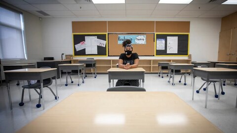 Une élève est seule dans une classe, assise à son bureau avec un masque.