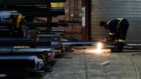 Devant des morceaux d'acier, un travailleur, penché, découpe une pièce et le processus fait des flammèches.
