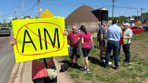Un groupe de personnes manifeste dehors. Une femme exhibe une pancarte jaune sur laquelle elle a écrit les lettres AIM avec une barre dessus.