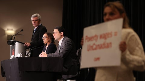 Air Canada officials speaking as a flight attendant holds a sign saying 'Poverty wages = UnCanadian'.