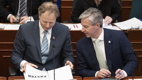 Allan MacMaster et Tim Houston sont assis à l'Assemblée législative. Ils baissent la tête vers des documents placés sur le bureau devant Allan MacMaster.