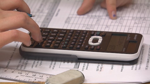 La main d'une femme sur une calculatrice. La calculatrice est posée sur des feuilles où l'on voit des colonnes de chiffres.