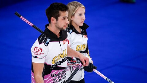 Une femme et un homme en uniforme, de profil, tiennent leurs balais de curling et regardent dans la même direction. 