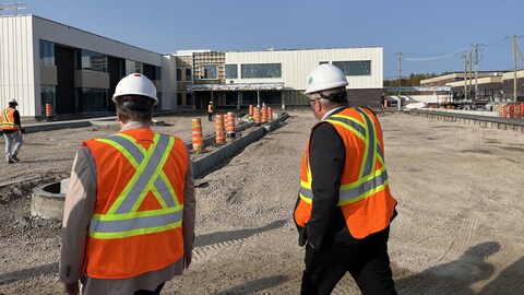 Deux personnes, vêtues de dossards et de casques, marchent sur un site de construction sur lequel se trouve une grosse bâtisse toujours en chantier. 