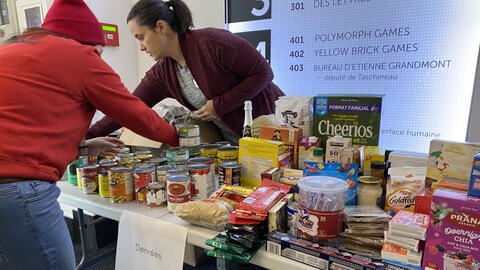 Deux personnes placent des boîtes d'aliments et des cannages sur une table. Le mot « denrées » est inscrit sur une feuille de papier, collée sur la table. 