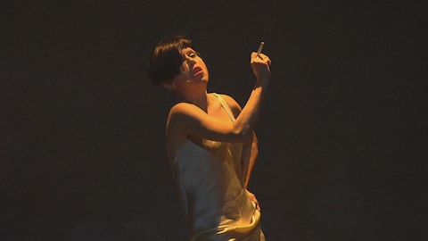 Une comédienne aux cheveux courts et vêtue d'une camisole blanche fume sur scène sous un éclairage sombre