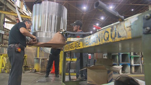 Des travailleurs manipulent une pièce mécanique suspendue dans une usine. 