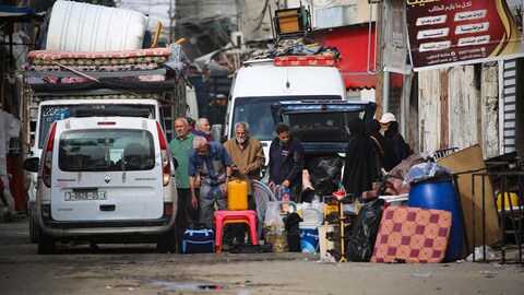 Une dizaine d'adultes sont debout dans une rue à côté de sacs, de tables et de chaises de plastique ainsi que de coussins sur le point d'être chargés dans un véhicule.