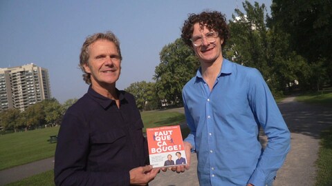 Deux hommes tiennent un livre, à l'extérieur, l'été. 