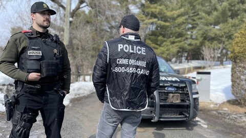 Deux policiers du crime organisé debout dans une entrée, devant un véhicule. 