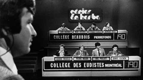 Dans un studio de télévision, l'animateur Michel Benoît fait face aux quatre participants du Collège Beaubois de Pierrefonds et à ceux du Collège des Eudistes de Montréal.
